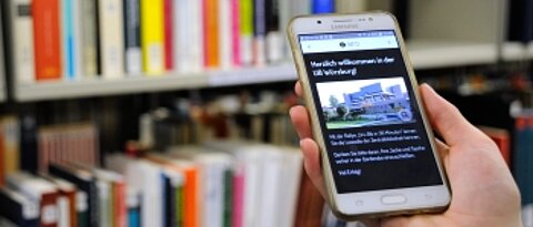 Eine Hand, die ein Smartphone hält. Auf dem Smartphone ist ein Ausschnitt der Actionboud-Führung UniBib in 30min zu sehen. Im Hintergrund ein Bücherregal